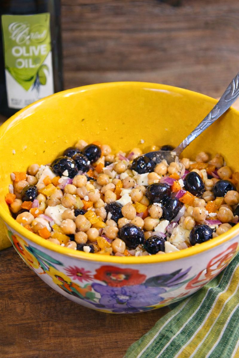 Mediterranean chickpea salad with black olives, feta cheese, red onion, and diced bell peppers served in a yellow bowl on a rustic wooden table with olive oil in the background.